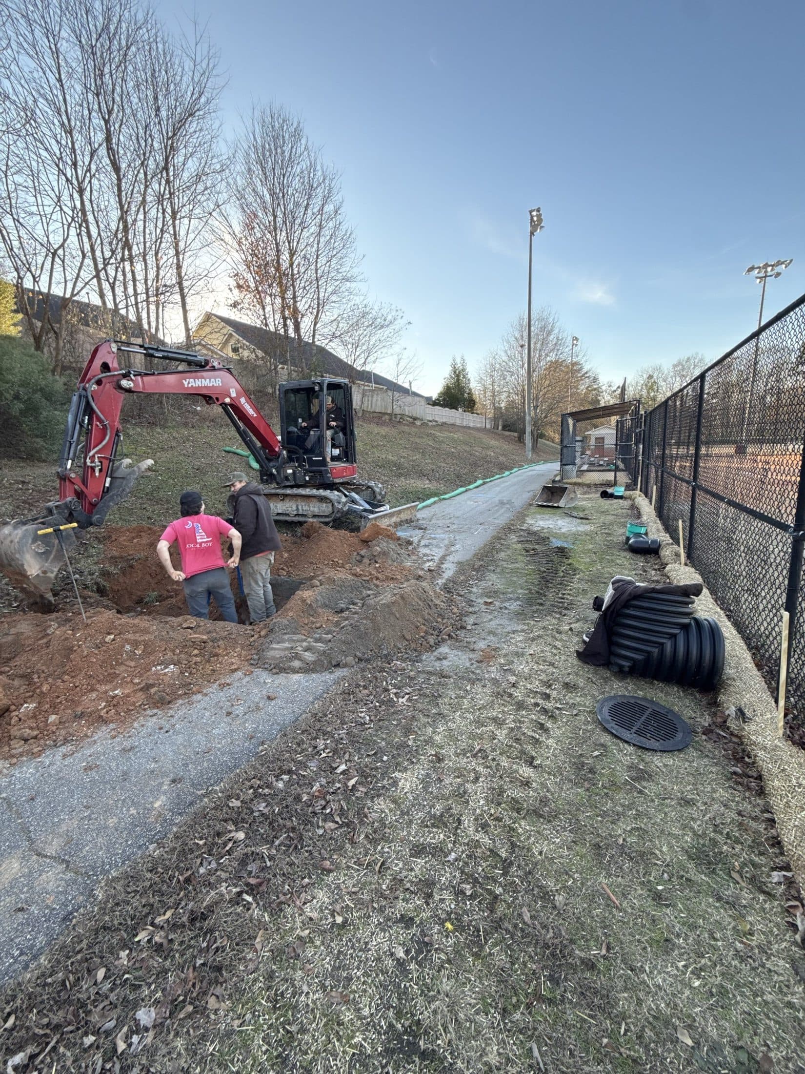 Crew setting a drainage structure beside the park trail with a mini excavator alongside the ball field fence