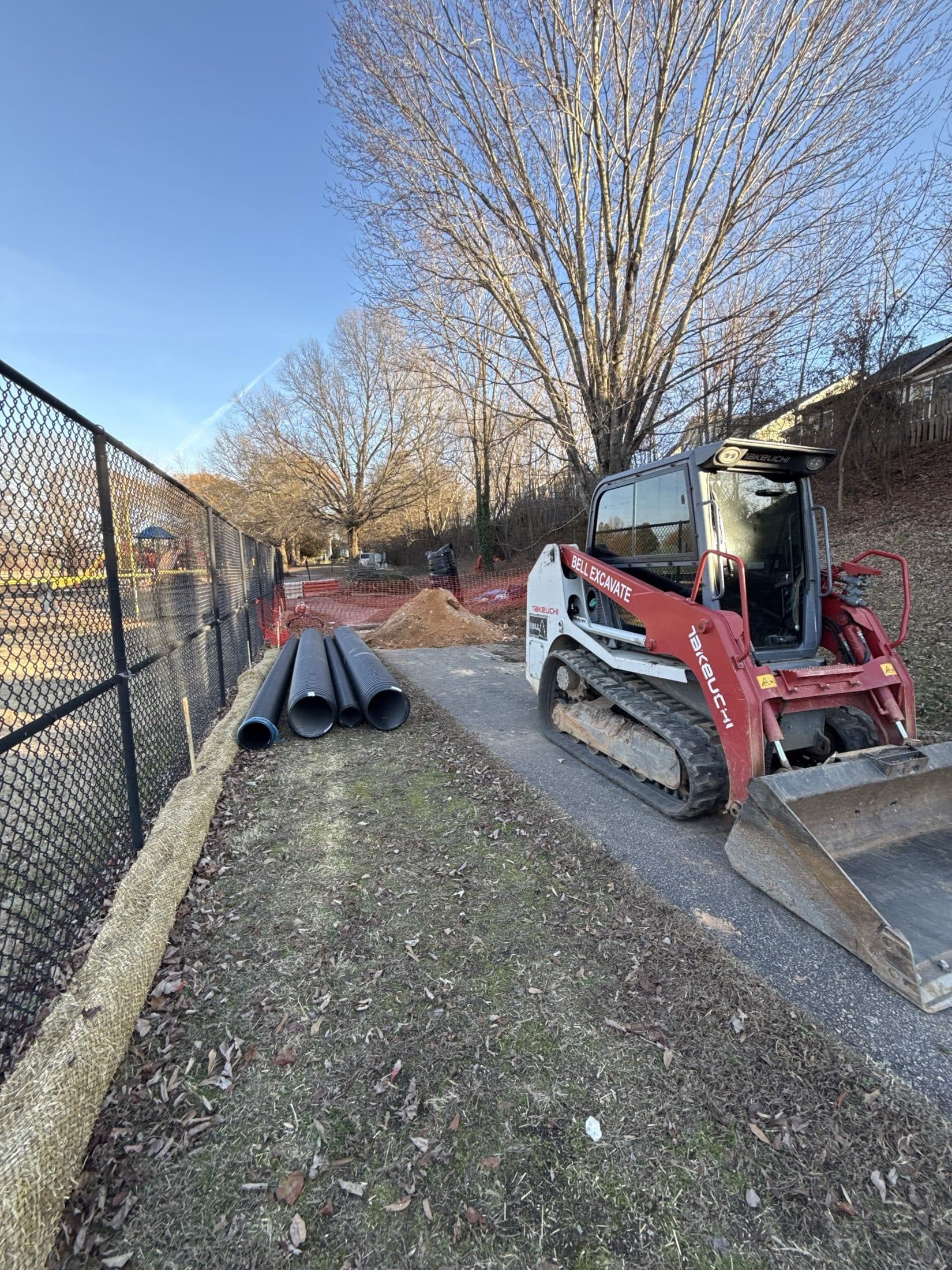 Track loader and stacked storm pipe staged along the trail corridor between the fenced fields