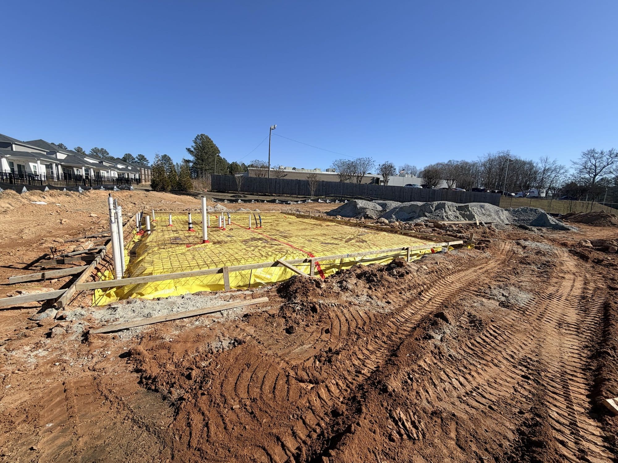 The prepared clubhouse slab from the site side, with earthwork and the community fence line beyond